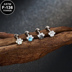 May include: Four titanium paw-shaped earrings with various colored gemstones. The earrings are silver-toned with a flat back and a paw print design. The gemstones include clear, blue, black, and iridescent colors. The text "ASTM F-136 TITANIUM" is in a white circle.