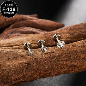 Puede incluir: Tres pequeños pendientes de joyería corporal plateados con parte posterior plana y tapas decorativas. Los pendientes están colocados sobre un trozo de madera marrón oscuro. La esquina superior izquierda de la imagen tiene un círculo blanco con el texto "ASTM F-136 TITANIUM".