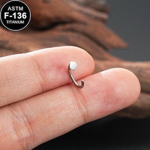Könnte beinhalten: Ein kleiner, gebogener Nasenring aus Titan. Der Ring hat eine silberne Oberfläche mit einer kleinen, flachen, weißen Scheibe oben. Der Text "ASTM F-136 TITANIUM" befindet sich in einem schwarzen Kreis oben links.