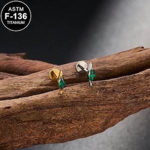 Könnte beinhalten: Zwei Schmuckstücke, eines goldfarben und eines silberfarben, jeweils mit einem grünen Edelstein. Das goldfarbene Stück hat einen flachen Rücken und einen spitz zulaufenden Edelstein. Das silberfarbene Stück hat einen spitz zulaufenden Edelstein. Der Text "ASTM F-136 TITANIUM" steht in einem Kreis.