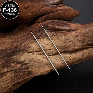 Puede incluir: Dos finas varillas de titanio plateado, posiblemente para piercing, se muestran sobre un fondo de madera oscura texturizada. Las varillas son rectas con puntas. Una etiqueta circular con el texto "ASTM F-136 TITANIUM" está en la esquina superior izquierda.