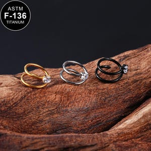 Puede incluir: Tres anillos de nariz con una sola piedra transparente en cada uno. Los anillos son de titanio y están disponibles en oro, plata y negro.