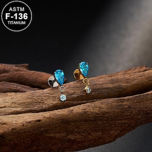 May include: A pair of earrings with teardrop-shaped, light blue gemstones. Each earring has a small, dangling, round, light blue gemstone. The earrings are set on a wooden surface. The text "ASTM F-136 TITANIUM" is in a white circle.