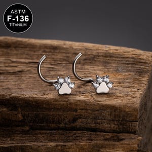 May include: Two silver nose rings with a paw print design and clear stones. The rings are made of ASTM F-136 titanium.
