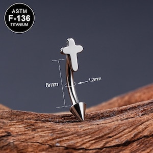 Puede incluir: Un piercing de cuerpo de cruz de titanio plateado con un extremo puntiagudo. El piercing mide 8 mm de largo y 1,2 mm de ancho. El texto "ASTM F-136 TITANIUM" es visible en la imagen.