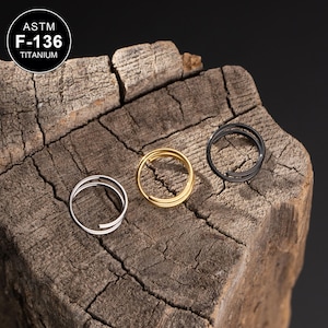 Może przedstawiać: Trzy tytanowe pierścienie w różnych kolorach: złoty, srebrny i czarny. Pierścienie są ułożone na kawałku drewna. Tekst "ASTM F-136 TITANIUM" jest widoczny w lewym górnym rogu obrazu.
