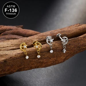 Puede incluir: Pendientes dorados y plateados con diseño de luna creciente y estrella, con una gema transparente colgante. Los pendientes se muestran sobre un trozo de madera. El texto "ASTM F-136 TITANIUM" está en un círculo en la esquina superior izquierda.