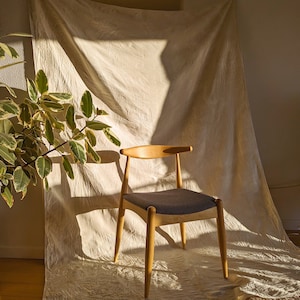May include: A wooden chair with a dark gray fabric seat sits on a white backdrop. The chair has a simple design with a curved back and four legs.