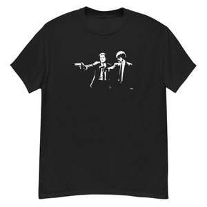以下が含まれることがあります： 黒いTシャツに、スーツを着た2人の男性が銃を構えている白いグラフィック。デザインはシルエットで、人物と武器が暗い生地に際立っています。Tシャツは快適な素材で作られています。