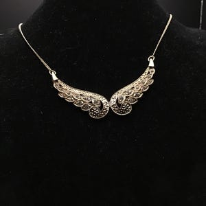 May include: A silver-toned necklace with a wing design. The necklace features two symmetrical wing-shaped pendants, each adorned with small, sparkling embellishments. The wings are connected by a delicate chain, creating an elegant and eye-catching accessory.