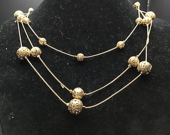 May include: Gold-toned necklace with three strands. Each strand features multiple filigree spheres of varying sizes, strung on delicate chains. The necklace is displayed against a black background, highlighting its intricate design and metallic sheen.