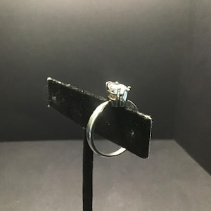 May include: A silver ring with a large, clear gemstone set on a black display stand. The ring's band is smooth and polished, and the gemstone is round and faceted, catching the light.