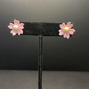 Puede incluir: Un par de pendientes de color rosa en forma de flor con un centro de perla. Los pendientes están en un expositor negro.