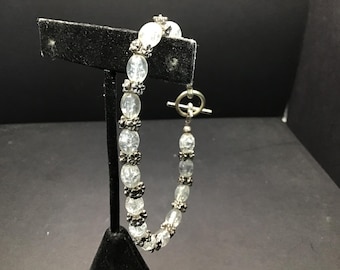 May include: A silver-toned bracelet featuring clear, oval-shaped beads interspersed with decorative silver accents. The bracelet has a toggle clasp and is displayed on a black stand, showcasing its design and craftsmanship.