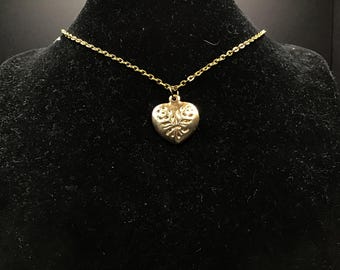 May include: A gold-colored necklace with a heart-shaped pendant. The pendant features a floral design and hangs from a delicate chain. The necklace is displayed on a black velvet surface.