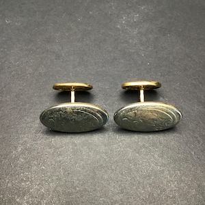May include: A pair of vintage cufflinks with an oval, silver-toned face and a gold-colored stem and backing. The silver face has a detailed, etched design. The cufflinks are displayed on a dark gray surface.