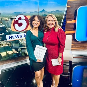 May include: Two women stand in front of a backdrop with a large "3" and the words "NEWS A". The woman on the left is wearing a green shirt and black skirt. The woman on the right is wearing a red dress. They are both holding books. The book in the woman on the left's hand has the title "Belly Binding: A Simple Perspective About Belly Binding for Your Body and Your Experience."