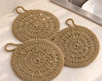 May include: Three circular, tan-coloured crocheted coasters or trivets, each featuring a central radial pattern and a small loop attached to the edge for hanging. The items are crafted from a textured, natural-toned yarn material.