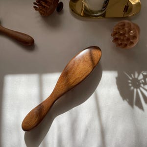 Puede incluir: Una herramienta de masaje de madera con un mango largo y una cabeza redondeada, junto con otras herramientas de masaje de madera. Las herramientas est&aacute;n hechas de madera c&aacute;lida de tono medio. Los art&iacute;culos est&aacute;n dispuestos sobre una superficie blanca.