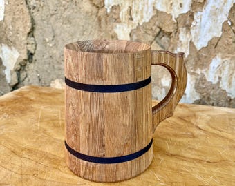 Könnte beinhalten: Ein handgefertigter Bierkrug aus Holz mit natürlicher Holzmaserung. Der Krug hat einen stabilen Griff und ist mit zwei schwarzen Bändern verziert. Der Krug wird auf einer Holzoberfläche präsentiert.