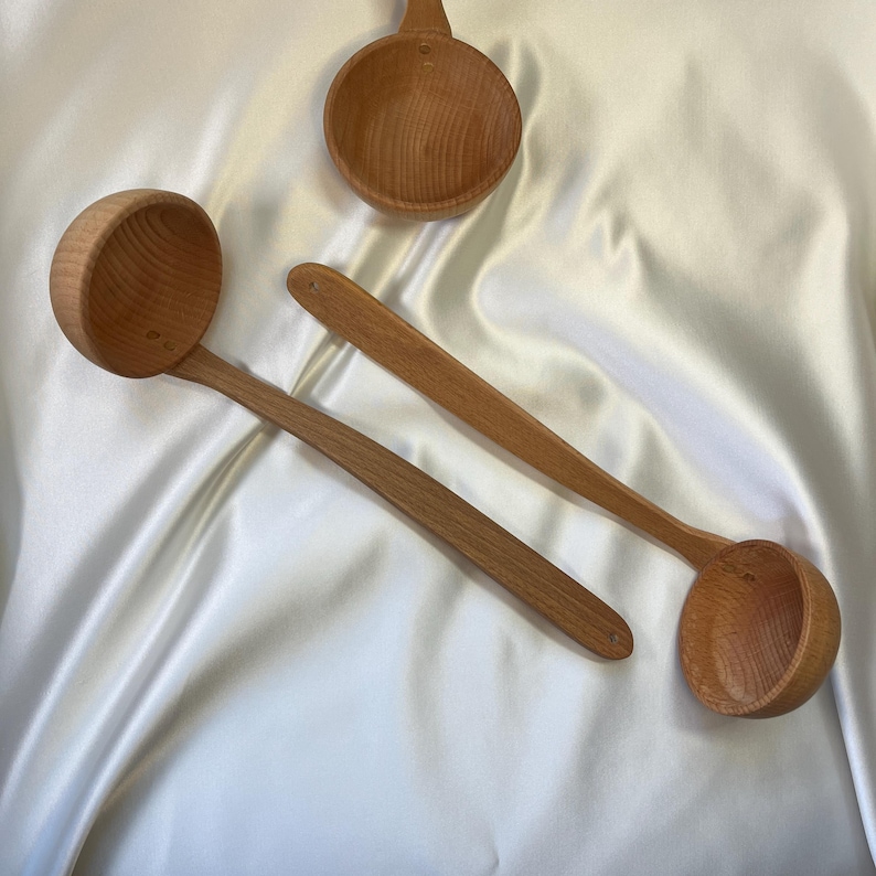 May include: Three wooden ladles and a small wooden bowl are arranged on a white satin surface. The ladles have long, slender handles and rounded bowls. The bowl is small and round with a short handle. The wood has a light brown color.