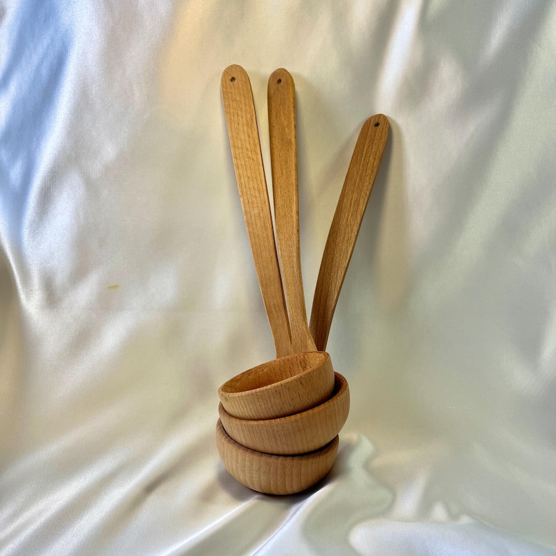May include: A set of four wooden ladles with long handles and rounded bowls. The ladles are made of light-colored wood and have a smooth finish. The bowls are stacked together, and the handles are fanned out.