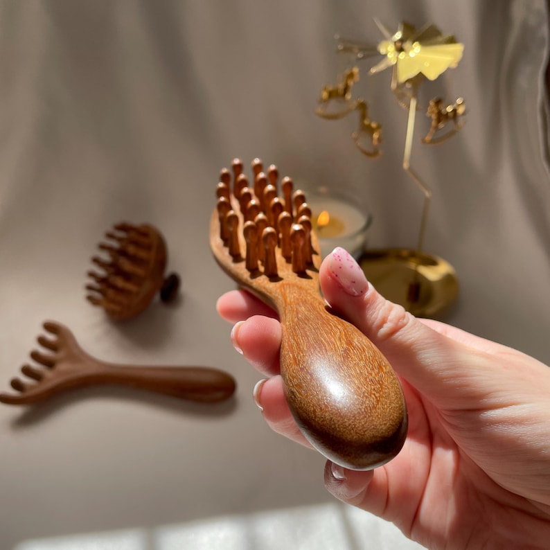 Puede incluir: Un masajeador de cuero cabelludo de madera con cerdas de madera redondeadas se sostiene en una mano. En el fondo hay otras dos herramientas de masaje de madera. Las herramientas est&aacute;n hechas de madera de color marr&oacute;n claro. El masajeador est&aacute; dise&ntilde;ado para la estimulaci&oacute;n y relajaci&oacute;n del cuero cabelludo.