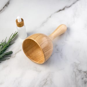 Peut inclure: Une pelle en bois de couleur claire avec un bol arrondi et un manche court, placée à côté d'un petit flacon compte-gouttes en verre dépoli avec un col en bambou et une poire en caoutchouc blanc, accompagnée d'un brin de romarin vert.