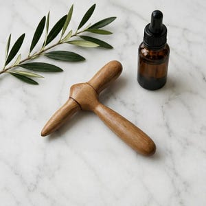 May include: A T-shaped wooden massage tool with a smooth, polished finish and a dark amber glass dropper bottle with a black cap, accompanied by a green olive branch with narrow leaves, all arranged on a white marble surface.