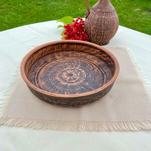 May include: A handmade, rustic ceramic bowl with a dark brown interior and a lighter brown rim. The bowl features a spiral design in the center and floral patterns around the edge. A small brown jug and red berries are in the background.