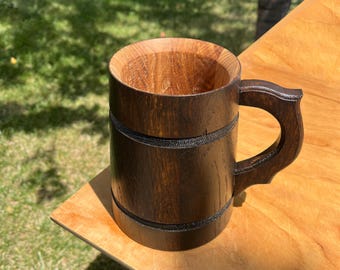 May include: A handcrafted wooden beer mug with a dark brown exterior and a lighter brown interior. The mug features a sturdy handle and is designed with a classic barrel shape. The mug is sitting on a wooden surface.