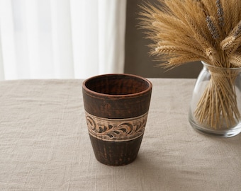 May include: A handmade, dark brown ceramic tumbler with a decorative band featuring a cream-colored, carved floral design. The tumbler is cylindrical and sits on a neutral-toned surface. A glass vase with wheat stalks is in the background.