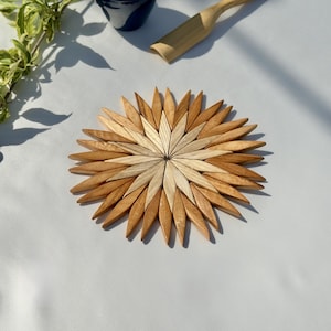 May include: A circular wooden trivet featuring a sunburst pattern composed of alternating light and medium-toned wood segments. The individual pieces are diamond-shaped and arranged in a radial design, creating a star-like appearance with a smooth finish.