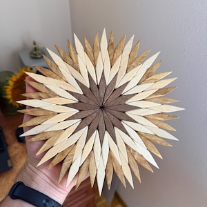 Op de afbeelding: Een ronde houten onderzetter met een sterpatroon bestaande uit talloze ruitvormige stukjes hout. Het ontwerp verloopt van een donkerbruin midden naar lichtere beige en crèmekleurige buitenlagen, wat een zonnestraaleffect creëert.