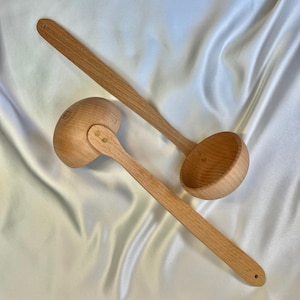 May include: Two wooden ladles with long handles and rounded bowls. The ladles are made of light-colored wood and have a smooth finish. One ladle is slightly smaller and has a hinged bowl. The background is a white, silky fabric.