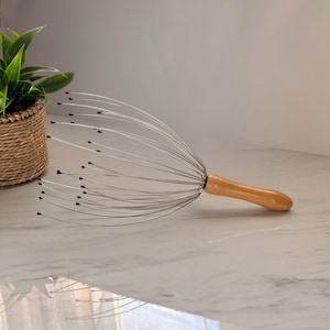 Puede incluir: Un masajeador de cabeza con un mango de madera y alambres metálicos flexibles que terminan en puntas negras. El masajeador está diseñado para estimular el cuero cabelludo. El mango es de color marrón claro y los alambres son plateados.