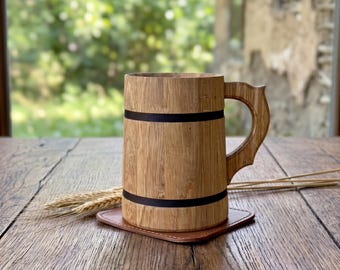 May include: A cylindrical wooden beer mug featuring a natural light brown wood grain, a curved handle, and two horizontal black bands encircling the body. The mug rests on a square brown leather coaster next to a small cluster of dried wheat stalks.