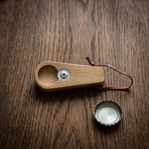 Peut inclure: Un ouvre-bouteille en bois avec une indentation circulaire et une vis métallique. Il est muni d'une boucle en cuir marron. Un bouchon de bouteille argenté est posé à proximité sur une surface en bois. L'ouvre-bouteille est conçu pour retirer les bouchons.