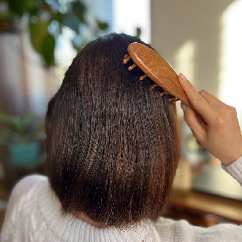 Puede incluir: Un cepillo de pelo de madera con dientes redondeados se utiliza para cepillar el cabello casta&ntilde;o oscuro. El cepillo es de madera clara y lo sostiene una persona que lleva un jersey blanco. El fondo est&aacute; borroso, con toques de vegetaci&oacute;n.