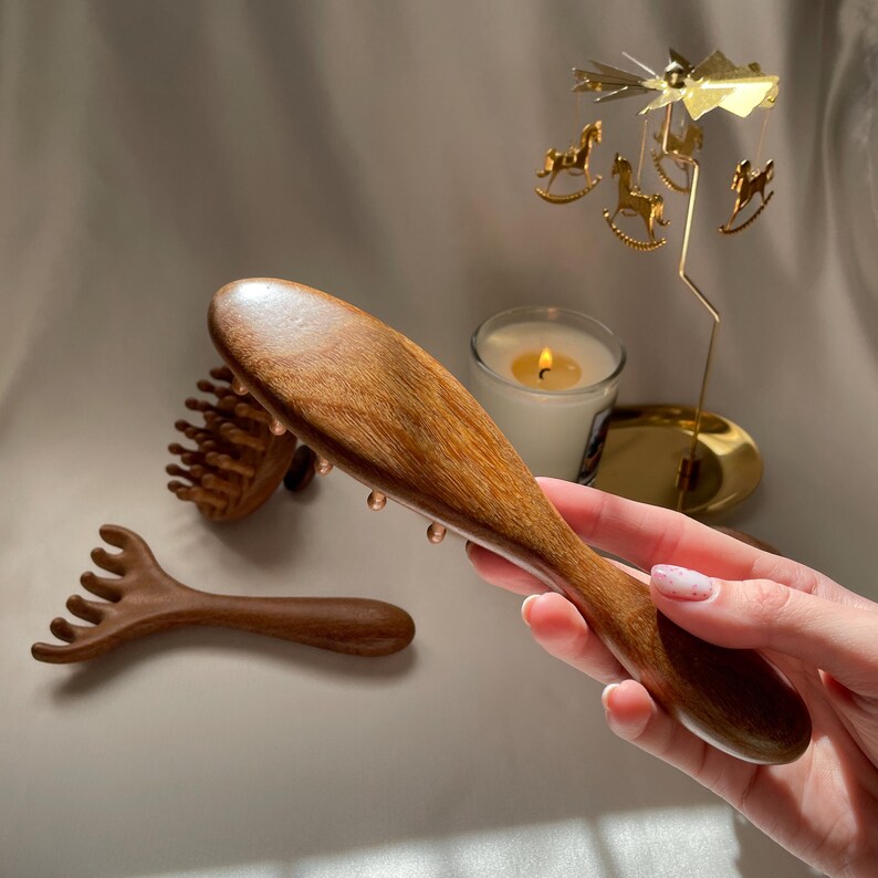 Puede incluir: Una herramienta de masaje de madera con un mango largo y una cabeza redondeada. En el fondo, hay otras dos herramientas de masaje de madera. Una vela y un carrusel dorado con caballos tambi&eacute;n est&aacute;n en el fondo.