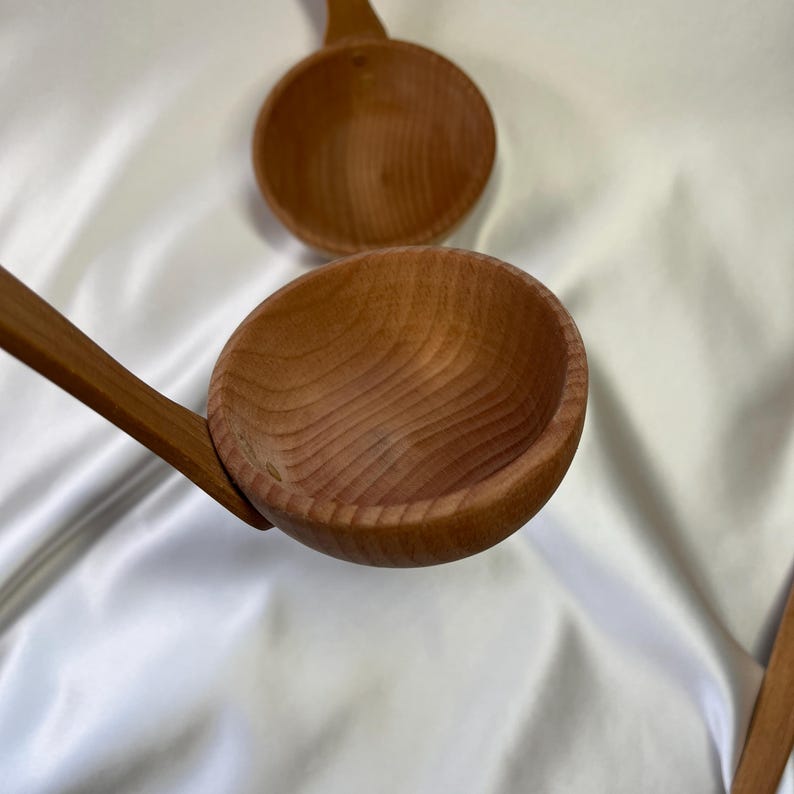 May include: Wooden ladle with a long handle and a round bowl. The ladle is made of light brown wood, with visible wood grain. The bowl is deep and smooth. Several ladles are visible, suggesting a set or multiple items.