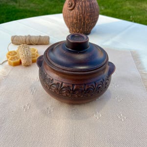 Może przedstawiać: Ciemnobrązowy ceramiczny garnek z pokrywką, z kwiatowym wzorem wokół podstawy. Garnek ma dwa małe uchwyty i stoi na beżowej tkaninie. Inne przedmioty to szpula sznurka, plaster miodu i dekoracyjny wazon w kształcie sowy.
