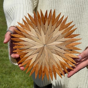 Può includere: Un sottopentola circolare in legno caratterizzato da un motivo a raggiera composto da pezzi di legno a forma di rombo, alternati in tonalità marrone chiaro e scuro, disposti in un design radiale con punte che si estendono dal centro.