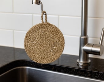 May include: A circular, beige, crocheted jute scrubby suspended from a silver tap. The scrubby has a loop for hanging and is positioned above a stainless steel sink. White tiles are in the background.