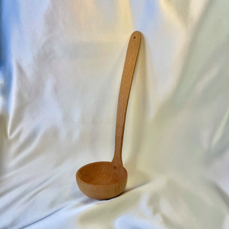 May include: A wooden ladle with a long, curved handle and a round bowl. The ladle is made of light-colored wood and has a small hole at the end of the handle. The bowl has a ribbed texture.