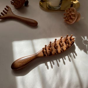 Puede incluir: Herramientas de masaje de madera, que incluyen un cepillo con clavijas redondeadas, un peine y un masajeador redondo. Las herramientas son de madera marr&oacute;n y est&aacute;n dispuestas sobre una superficie blanca, lo que sugiere un tema de spa o bienestar.