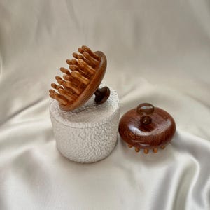 Op de afbeelding: Twee houten hoofdhuidmassagers. Eén is een ronde, handheld massager met meerdere afgeronde tanden. De andere is een kleinere, ronde massager met een handvat. Beide zijn gemaakt van warm, roodbruin hout en liggen op een wit, getextureerd oppervlak.