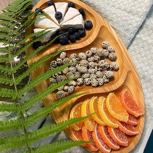 May include: A wooden serving tray with three sections. The top section has a wedge of brie cheese and blueberries. The middle section has sesame-coated candies. The bottom section has orange and pink fruit slices.