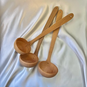 May include: Four wooden ladles with long handles and rounded bowls are arranged on a white, silky surface. The ladles are made of light-colored wood, with a natural finish. Each ladle has a small hole at the end of the handle.