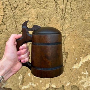 Peut inclure: Une chope à bière en bois brun foncé avec un couvercle et une poignée. La poignée est munie d'un ouvre-bouteille.